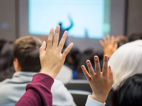The back of two students' raised hands in a classroom setting, other seated students and an instructor in front of a lit screen in the background